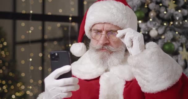 Gros plan portrait d'un Père Noël en pleine couvée lit le message sur son smartphone, à travers ses lunettes abaissées, hochant la tête avec une décision négative, avec l'incapacité de satisfaire un souhait des enfants. — Video