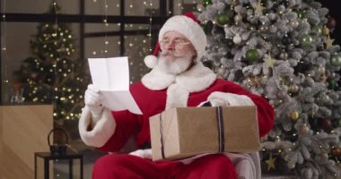 Noel Baba karnavalı kostümlü neşeli büyükbaba, Noel ağacı arka planında kocaman bir Noel hediyesi kutusuyla koltukta otururken çocuk mektupları okuyor.