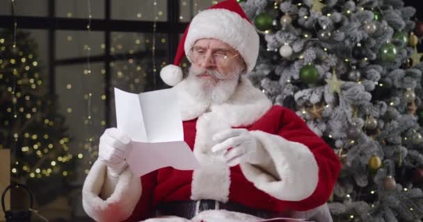 Front view of cheerful Santa Claus wearing Christmas costume with red hat, sitting in armchair against Christmas tree at his residence, reading letter, then smiling toothy smile looking at camera — Stock Video