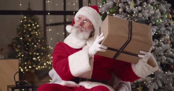 Ένας ηλικιωμένος καλός παππούς ντυμένος Άγιος Βασίλης στην κατοικία του στο φόντο ενός διακοσμημένου χριστουγεννιάτικου δέντρου και φέρνει ένα χριστουγεννιάτικο δώρο στο αυτί του, κουνώντας το υπενθυμίζοντας τι βρίσκεται εκεί — Αρχείο Βίντεο