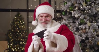 Noel Baba karnavalında gri sakallı neşeli, yaşlı beyaz bir adam. Kırmızı ve beyaz bir kostüm. Şapkası Noel ağacının dibinde oturuyor ve akıllı telefonuyla bilgisayar oyunları oynarken eğleniyor.