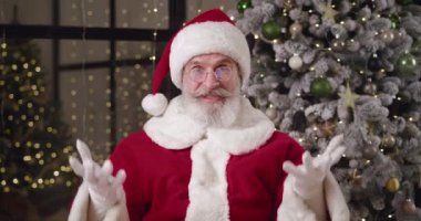 Noel Baba kostümü giymiş neşeli yaşlı bir adam evde süslü bir Noel ağacına karşı kameranın önünde oturur ve kameraya çekilirken el sallar. Çevrimiçi kavram