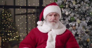 Mutlu, iyi huylu, yaşlı bir Noel Baba 'nın Noel ağacı arkaplanına karşı evde oturmasının ön görüntüsü, önündeki web kamerasına dikkatle bakıyor.