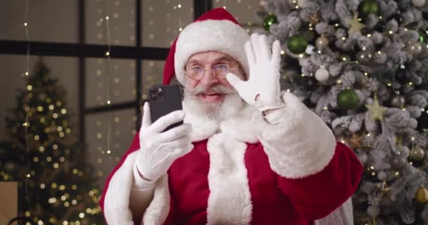 Dobromyslný starší Santa Claus ve své rezidenci, sedí na křesle u krásného vánočního stromku a mluví videohovory. Santa Claus komunikace on-line pomocí moderních bezdrátových technologií — Stock video