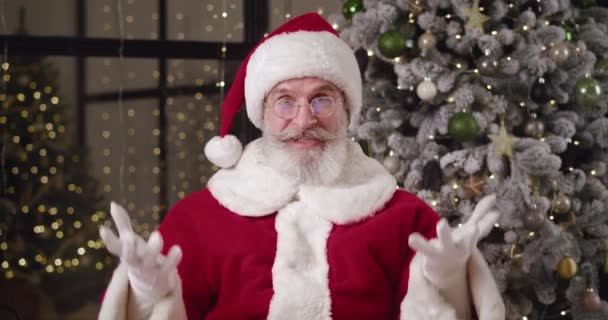 Cheerful elderly man in Santa Claus costume sits in front of a web camera against a decorated beautiful Christmas tree at home and waves his interlocutor while taking by video call. Online concept — Stock Video
