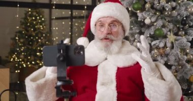 Evindeki neşeli, yakışıklı Noel Baba 'nın yakın çekimi, çocuklardan Noel hediyesi almak, tripoda monte edilmiş akıllı bir telefonun önünde otururken görüntülü konuşma yapmak.