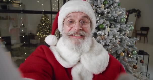 Close-up of a cheerful Santa Claus holding a mobile phone in his outstretched hand, making a video call, waving hello with hand while greeting his interlocutor standing against Christmas tree at home — Stock Video