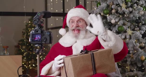 Snäll jultomte inspelning video vädjan till barn som innehar julklapp låda, sitter framför en digitalkamera monterad på ett stativ, mot bakgrund av en julgran i hans bostad — Stockvideo