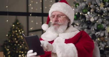 Büyüleyici yaşlı Noel Baba, doğal gri sakallı ve not defterindeki notları okuyarak geleneksel kutlamalar sırasında evinde yanan Noel ağacı arka planında oturuyor.