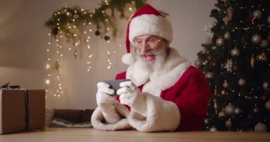Karnaval kırmızı beyaz takımlı neşeli yaşlı Noel Baba bir Noel ağacının arka planında oturur ve eğlenir, akıllı telefonda bilgisayar oyunları oynar, kazanırken yumruğunu sıkar.