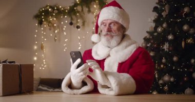 Noel Baba akıllı telefon kullanıyor, ışıklandırılmış bir Noel ağacına karşı oturuyor, gülümsüyor, gülüyor, seviniyor ve internette harika haberler alırken yumruklarını sıkıyor. Kablosuz teknoloji ve Noel konsepti