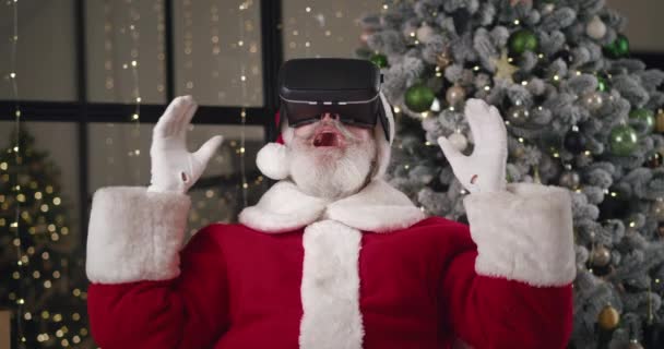 Curious elderly charming Caucasian man in Santa Claus carnival costume puts on VR glasses and plunges into the world of virtual reality. Santa moves his hands while surfing the virtual reality — Stock Video