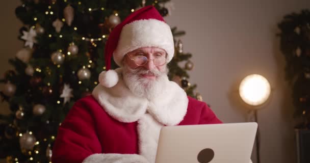 Χαρούμενος ηλικιωμένος γενειοφόρος Άγιος Βασίλης κάθεται σε ένα τραπέζι, πίνοντας ζεστό ρόφημα, ενώ εργάζονται σε φορητό υπολογιστή κατά ένα φωτισμένο φόντο χριστουγεννιάτικο δέντρο στη ζεστή χειμερινή κατοικία του. Χριστουγεννιάτικες διακοπές — Αρχείο Βίντεο
