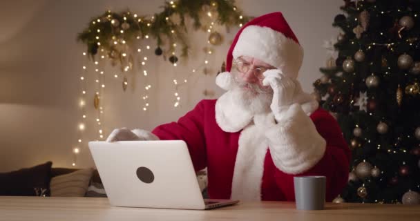 Miły, starszy Święty Mikołaj zdejmuje okulary po długiej pracy online w domu, zamyka pokrywę laptopa i zasypia na niej, siedząc na tle migoczących światełek choinki — Wideo stockowe