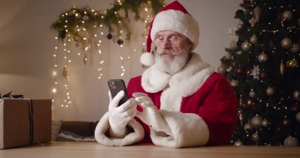Santa Claus menggunakan smartphone, duduk di belakang pohon Natal menyala, tersenyum, tertawa, sukacita dan kepalan tangan ketika menerima berita besar secara online. Teknologi nirkabel dan konsep Natal — Stok Video