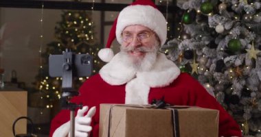 Evindeki yakışıklı Noel Baba 'nın portresi, elinde büyük bir Noel hediyesi kutusuyla Noel ağacının arka planındaki tripoya monte edilmiş akıllı bir telefonun önünde duruyor.