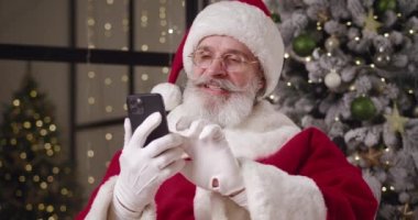 Neşeli bir Noel Baba web sitelerini karıştırıyor, internette geziniyor, akıllı telefonlar kullanıyor, neşeli, gülümseyen, yumruklarını sıkan, modern akıllı telefonundan harika haberler okuyor. Noel Baba online