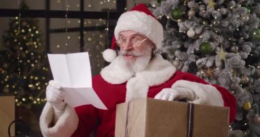 Çekici Noel Baba 'nın elinde bir yeni yıl hediyesi kutusu, başını sallıyor, Noel hediyesi kutusundan, içinde çocukların dilekleri olan bir kağıt parçasına bakıyor, cömert bir gülümsemeye doğru yayılıyor.