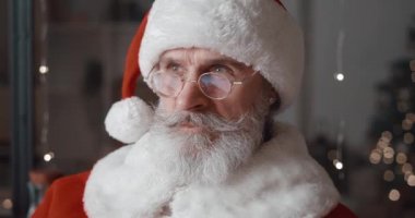 Noel Baba karnavalında sakallı yakışıklı bir adamın özgüvenli portresi. Karnaval kıyafeti ve gözlük takıyor. Düşünceli bir şekilde kameraya dönüp neşeli bir gülümsemeyle gülümsüyor.