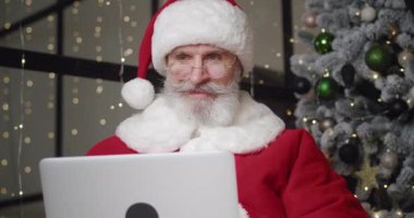 Noel Baba kostümlü olgun sakallı bir adam bir Noel ağacının arka planında oturuyor ve dizüstü bilgisayarında klavye yazıyor çocukların dileklerini içeren e-postaları okuyor.