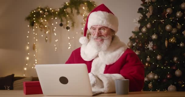 Homem idoso barbudo vestido de Papai Noel sentado à mesa com laptop e conversando em videochamada, mostrando o polegar até a câmera da web e uma caixa de presente vermelha, contra a árvore de Natal iluminada e cenário de decorações — Vídeo de Stock