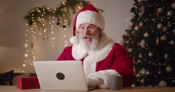 Веселий старий Санта Клаус із добродушною вдачею відпочиває у своєму домі, посміхається, радіє, стискає кулаки, читаючи чудові новини на ноутбуці, сидячи навпроти різдвяного дерева. — стокове відео