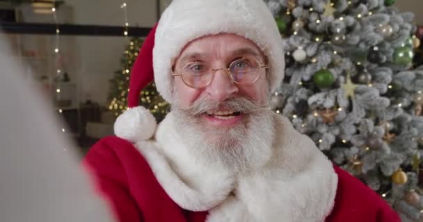 Όμορφος γενειοφόρος ηλικιωμένος άνδρας ντυμένος ως Άγιος Βασίλης μιλάει με βιντεοκλήση, επικοινωνεί online κρατώντας το κινητό του τηλέφωνο σε τεντωμένα χέρια ενάντια στο χριστουγεννιάτικο δέντρο κατά τη διάρκεια των γιορτών των Χριστουγέννων — Αρχείο Βίντεο