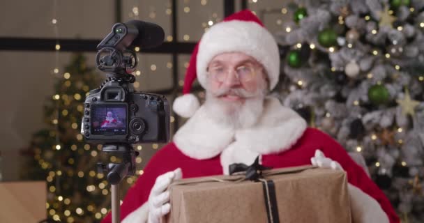 Εστίαση στην ψηφιακή κάμερα τοποθετημένη στο τρίποδο σε θολή φόντο ενός χαρούμενου Αϊ Βασίλη που καταγράφει μια έκκληση βίντεο για τα παιδιά, λαμβάνοντας παραγγελίες για δώρα και gesturing κρατώντας τεράστια χριστουγεννιάτικο κουτί δώρου — Αρχείο Βίντεο