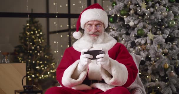 Playful modern Caucasian man with gray beard in Santa Claus carnival red and white costume with hat sitting against a Christmas tree background and enjoying playing computer games on his smartphone — Stock Video