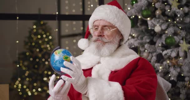 Papai Noel senta-se por uma árvore de Natal iluminada em sua residência, segura um globo e vira-lo, olha atentamente para os continentes, escolhendo as instruções para ir, em seguida, aponta o dedo no ponto no mapa — Vídeo de Stock