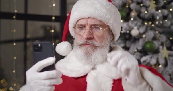 Κοντινό πλάνο του σύγχρονου Αϊ Βασίλη στην κατοικία του με φόντο το χριστουγεννιάτικο δέντρο, διαβάζοντας το μήνυμα στο smartphone του μέσω χαμηλωμένα γυαλιά του και χαίρεται σφίγγοντας γροθιές. Νέο Έτος έννοια — Αρχείο Βίντεο