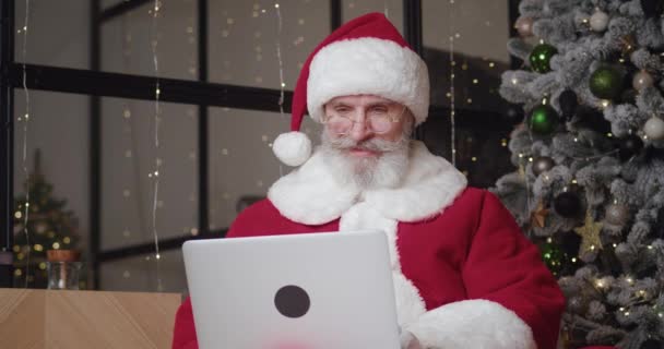 Κοντινό πλάνο. Καλός-natured ηλικιωμένος άνδρας στο κοστούμι καρναβάλι Άγιος Βασίλης κάθεται σε μια πολυθρόνα στο σαλόνι κοντά στο χριστουγεννιάτικο δέντρο και πληκτρολογώντας στο πληκτρολόγιο laptop, διαβάζοντας μηνύματα ηλεκτρονικού ταχυδρομείου με τα παιδιά επιθυμεί — Αρχείο Βίντεο