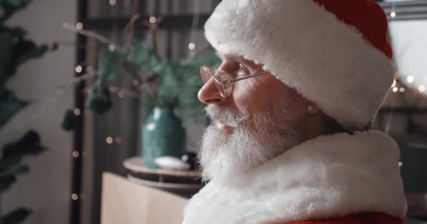Boczne zbliżenie wesołego brodatego staruszka w stroju karnawałowym Świętego Mikołaja patrzącego prosto przed siebie, a następnie uśmiechającego się szczęśliwie do kamery, siedzącego w domu na tle choinki — Wideo stockowe