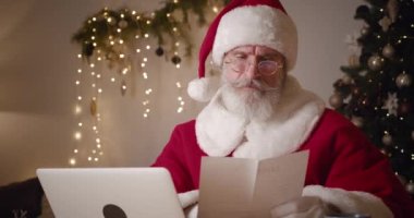 Yaşlı Noel Baba bir çocuk mektubu açar ve dizüstü bilgisayarda çalışır, tatlı tatlı başını sallayarak kışın süslediği yanan bir Noel ağacının arka planında oturur.