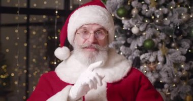 İyi huylu, yakışıklı Noel Baba işaret parmağını dudaklarına koyar, dişlek bir gülümsemeyle kameraya bakar, rahat evindeki Noel ağacının karşısında durur.