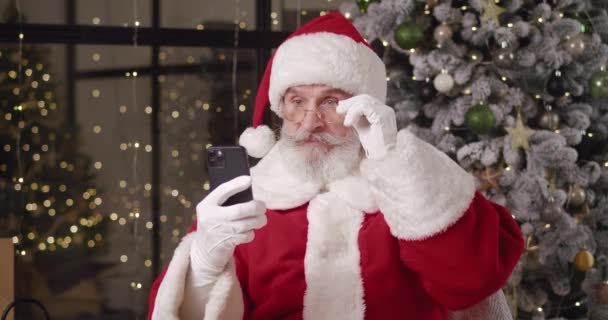 Kedves érett Mikulás örül és nevet olvasás közben üzenetet mobiltelefonján, mosolyog fogas mosoly, és ökölbe szorítja a boldogság, hogy képes kielégíteni a gyerekek kívánságait karácsonyi ajándékok — Stock videók