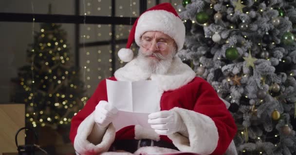 Papá Noel en traje de Navidad sentado en sillón contra el árbol de Navidad en su residencia, leyendo cuidadosamente la carta de los niños, muestra un pulgar hacia arriba y luego sonríe con alegre sonrisa dentada — Vídeos de Stock