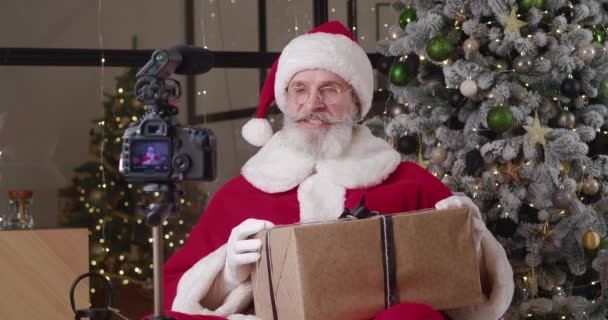 Άγιος Βασίλης καταγραφή βίντεο έκκληση σε παιδιά που κατέχουν χριστουγεννιάτικο κουτί δώρου, κάθεται μπροστά από θολή ψηφιακή φωτογραφική μηχανή τοποθετημένη σε ένα τρίποδο, στο φόντο ενός χριστουγεννιάτικου δέντρου στην κατοικία του — Αρχείο Βίντεο