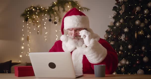 Основное внимание уделяется негативным эмоциям и выражениям лица Санта-Клауса, смотрящего на монитор ноутбука через опущенные очки, чувствующего разочарование и недовольство, читающего плохие новости на Рождество — стоковое видео
