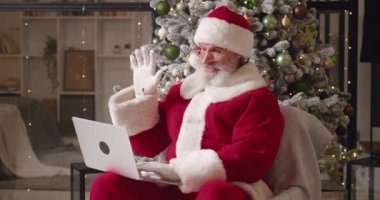 Evinde, arkasında bir Noel ağacı olan neşeli Noel Baba, dizüstü bilgisayar kullanıyor, video görüşmesi sırasında selam veriyor ve başparmağını kaldırıyor. Noel Baba, geleneksel Noel etkinliğinde internette sohbet ediyor.