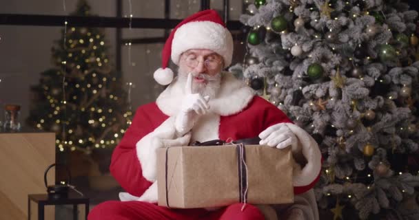 Веселый Санта-Клаус с новогодним подарком на фоне красивой елки, прикладывая указательный палец к губам и развязывая бантик на подарок, улыбаясь, глядя в камеру — стоковое видео