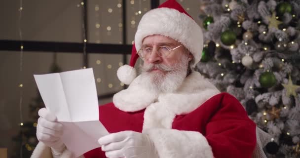 Καλαίσθητο είδος Ο Άγιος Βασίλης χαϊδεύει το μουστάκι και τη γενειάδα του, διαβάζοντας προσεκτικά παιδικά γράμματα με επιθυμητά δώρα, κουνάει το κεφάλι του, δείχνοντας τον αντίχειρά του και απλώνεται σε ένα γενναιόδωρο χαμόγελο. — Αρχείο Βίντεο