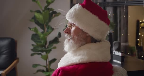 Retrato de cerca de un Santa Claus pensativo mirando pensativamente delante de él, luego a la cámara a través de sus gafas bajadas y extendiéndose en una hermosa sonrisa de dientes alegres. — Vídeo de stock