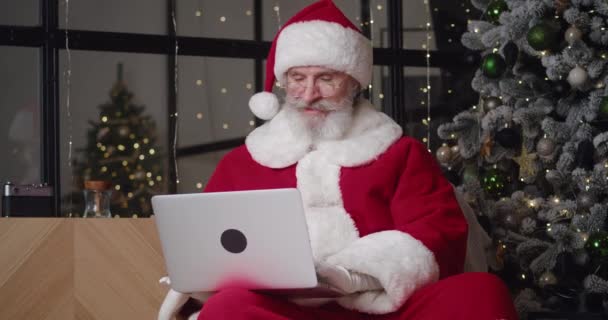 Handsome good-natured elderly man in Santa Claus carnival costume sitting on an armchair in the living room near Christmas tree and typing on laptop keyboard, reading emails with the children wishes — Stock Video