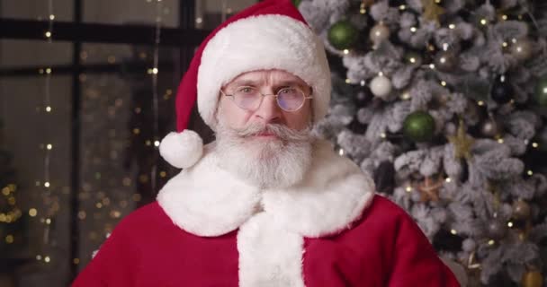 Απογοητευμένος και απογοητευμένος, ο Άγιος Βασίλης κοιτάζει την κάμερα μέσα από τα χαμηλωμένα γυαλιά του και αφήνει ένα χαρούμενο χαμόγελο καθώς στέκεται δίπλα στο χριστουγεννιάτικο δέντρο στην όμορφη κατοικία του.. — Αρχείο Βίντεο