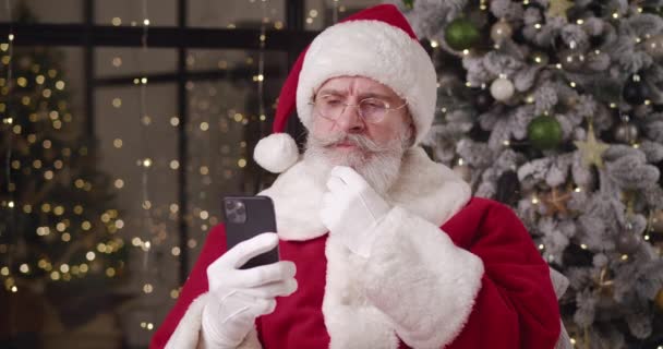 Задумчивый Санта читает сообщение на своем смартфоне, гладит усы и оглядывается вокруг, кивая головой с отрицательным решением, с неспособностью удовлетворить детское желание. — стоковое видео