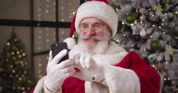 Κοντινό πλάνο του Άγιου Βασίλη που κάθεται απέναντι στο χριστουγεννιάτικο δέντρο στην κατοικία του, χρησιμοποιώντας το κινητό τηλέφωνο, κλέβοντας, διαβάζοντας ειδήσεις, χαλαρωτική περιήγηση σε ιστοσελίδες, κρατήσεις, online αγορές. Άγιος Βασίλης που χρησιμοποιεί internet — Αρχείο Βίντεο