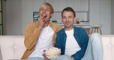 Gülümseyen erkek eşcinsel çift evde vakit geçiriyor. Kanepede dinleniyorlar ve evde patlamış mısırla televizyonda komedi filmi izliyorlar.