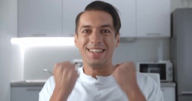 Başarıyı kutlayan, kameraya şaşıran, evdeki oturma odasındaki kanepede oturan genç adamın yakınlaşması. Zafer konsepti. Evet kazanan jest.