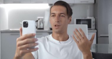 Akıllı telefon tutan genç bir adam oturma odasında kanepede otururken selfie videosu çekiyor. Gülümseyen adamın portresi mobil video arıyor, evdeki kanepede dinleniyor.